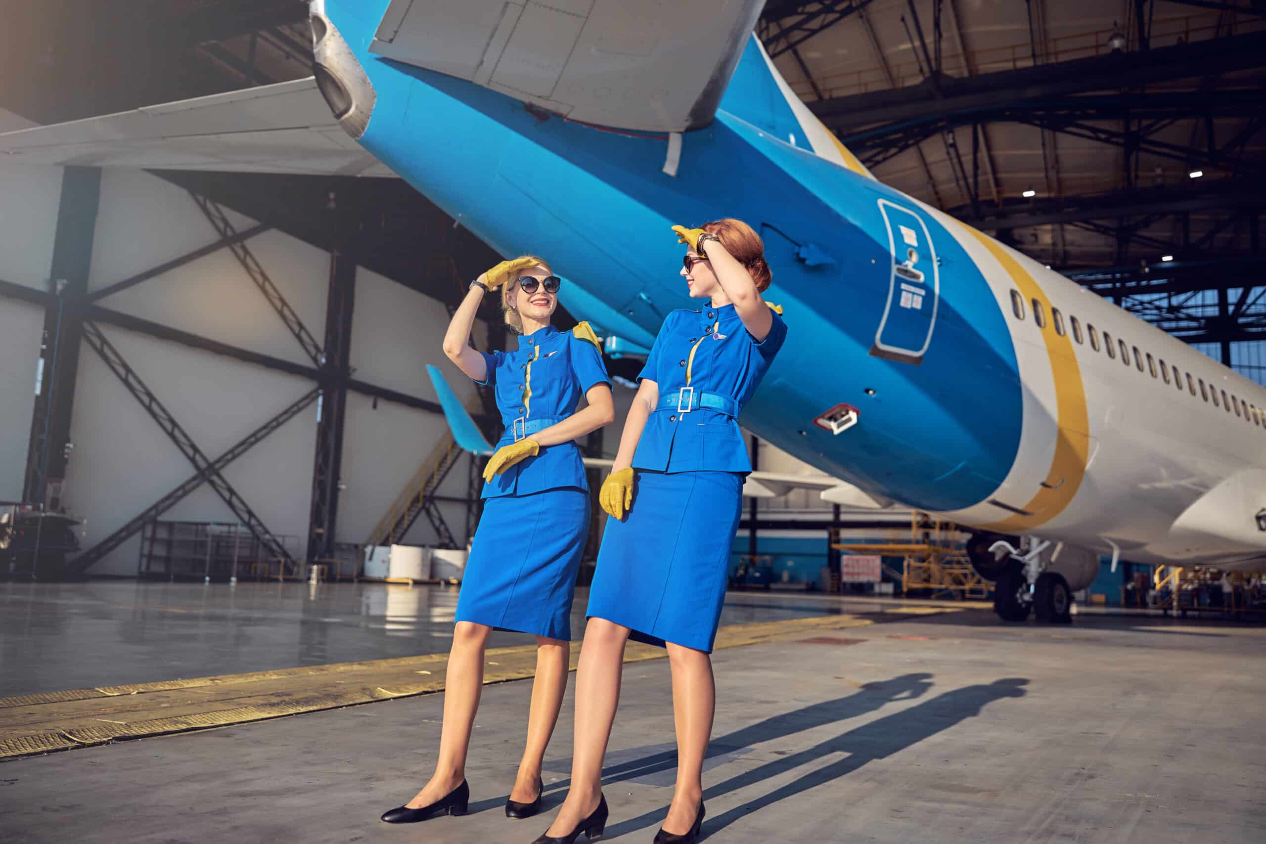 Full length portrait of elegant flight attendant wearing blue respective uniform holding hand near head while looking to each other in the aviation hangar